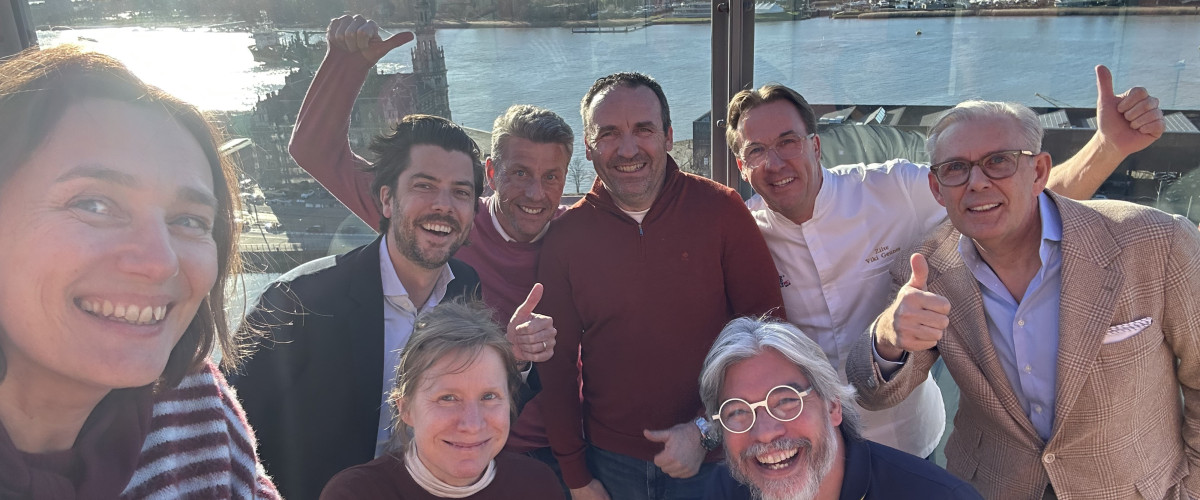 Groepsselfie op dakterras met uitzicht Groepsselfie van zeven mensen op een dakterras met duimpjes omhoog, met rivier en stadsgezicht op de achtergrond.